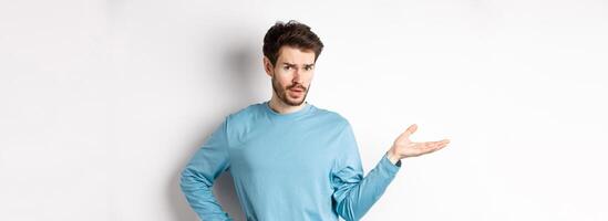 So what. Confused young man raising hand and looking skeptical at camera, know nothing, cant understand something, standing on white background photo