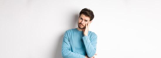 Sad young man looking frustrated, touching temple of head and frowning uneasy, standing against white background photo