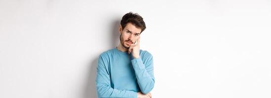 Skeptical young man listening to you with bored face, looking at camera reluctant, standing in sweatshirt over white background photo
