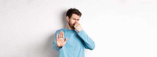 Image of caucasian man stretch out hands to stop or reject something, shut nose disgust, refusing product with bad smell, standing over white background photo