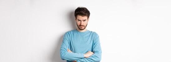 Image of offended and sad young man with beard, look from under forehead and sulking irritated, cross arms on chest defensive, mad at someone, standing on white background photo