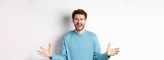 Image of frustrated and worried young man shaking hands, trying to explain something, standing anxious against white background photo