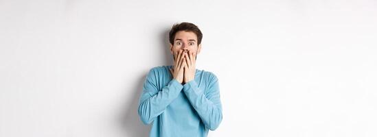 Shocked and startled cauacasian man, gasping and covering mouth with hands, staring at something scary, standing over white background photo