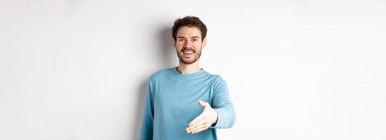 Friendly smiling man stretch out hand, saying hello and give arm for handshake, standing over white background photo