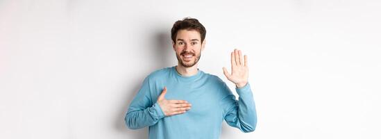 Smiling hosent man raising arm and hold hand on heart, making promise tell truth, swear or give oath, standing over white background photo