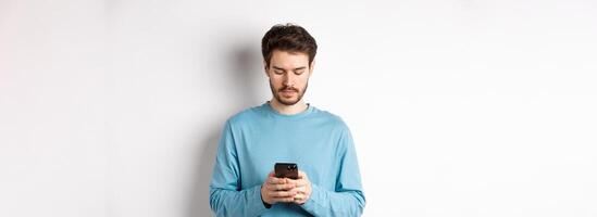 Serious young man reading message on smartphone, looking at mobile screen, standing over white background photo