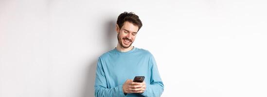 Portrait of modern caucasian man with beard, wearing casual clothes, reading smartphone screen and smiling, networking via mobile phone, standing over white background photo