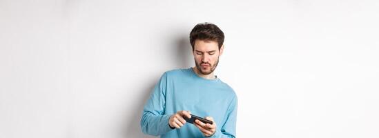 Young man playing games on smartphone, tilt body and tap mobile screen, standing joyful over white background photo