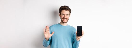 Handsome caucasian man with beard, showing empty smartphone screen and number five, raising hand to wave and say hello, standing on white background photo