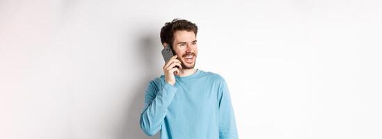 Technology concept. Joyful man enjoying phone call, talking on smartphone and smiling, standing in casual shirt over white background photo
