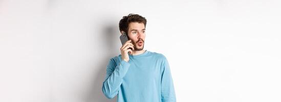 Technology concept. Intrigued young man looking left impressed, talking on mobile phone, calling someone, standing over white background photo