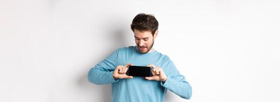 E-commerce and shopping concept. Caucasian man showing blank mobile screen and looking at it, standing curious against white background photo