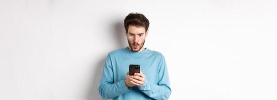 Amazed young man staring at smartphone screen and saying wow, looking at online offer, standing on white background photo