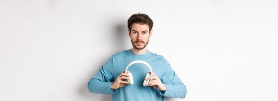 Young bearded guy in blue sweatshirt put on wireless headphones, listening music, standing on white background photo