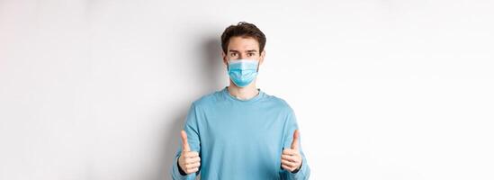 Covid-19, pandemic and social distancing concept. Young man in casual clothes and medical mask, showing thumbs up and looking serious, protecting from virus during quarantine photo