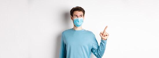 Coronavirus, health and quarantine concept. Man in medical mask smiling pleased, looking and pointing finger left at logo, showing promo offer, white background photo
