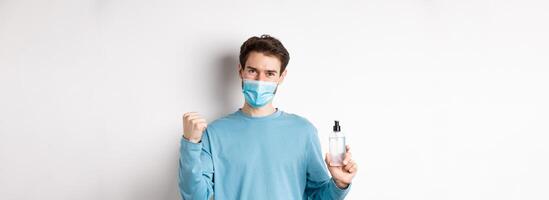 Covid-19, health and quarantine concept. Cheerful man in face mask celebrating, showing fist pump and bottle with hand sanitizer, fighting germs, white background photo