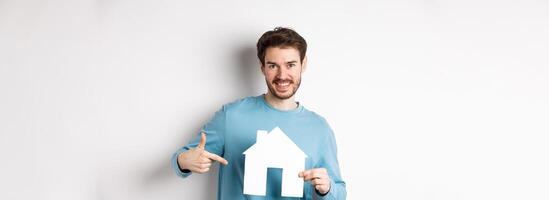 Real estate and insurance concept. Smiling young man pointing at paper house cutout and looking happy, buy apartment, standing over white background photo