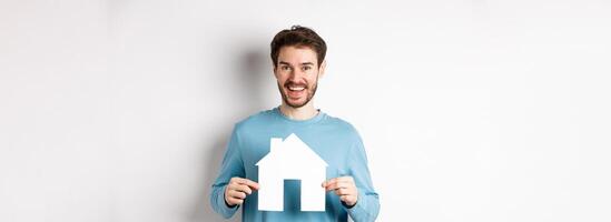 Real estate and insurance concept. Handsome modern man buying property, smiling and showing paper house cutout, standing over white background photo