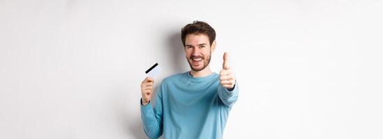 Cheerful young man showing plastic credit card and thumb up, like and approve good bank service, standing over white background photo