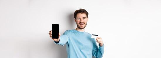 E-commerce and shopping concept. Smiling caucasian man showing plastic credit card and empty smartphone screen, recommending online app, white background photo