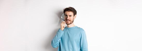 Smiling young man detective looking through magnifying glass, searching for promo offer, standing on white background photo