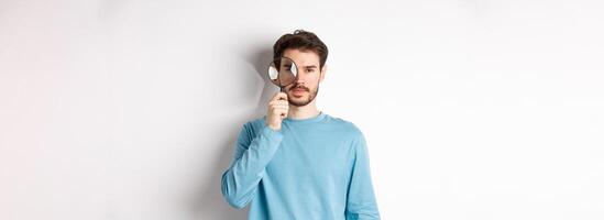 Man searching for something, looking through magnifying glass at camera, standing on white background photo