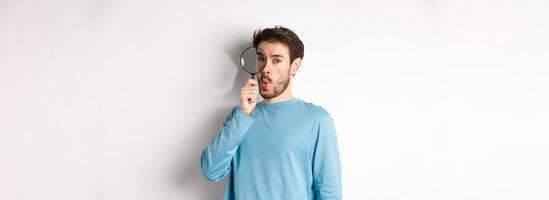 Surprised man found something, looking through magnifying glass with amazed face, standing over white background photo
