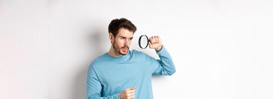 Young handsome man look through magnifying glass with curious face, investigating or searching for something, standing on white background photo