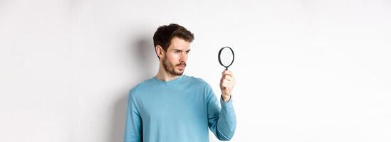 Young handsome man look through magnifying glass with curious face, investigating or searching for something, standing on white background photo