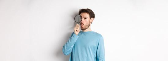 Curious guy looking through magnifying glass on right logo, saying wow, found cool thing, standing on white background photo
