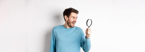 Handsome smiling man looking through magnifying glass with amazed face, found interesting promotion, standing on white background photo