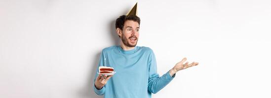 Celebration and holidays concept. Surprised man shrugging in disbelief, looking away, holding birthday cake and wearing party cone hat, white background photo