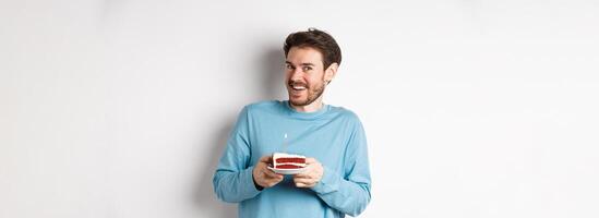 Excited man receive birthday surprise, holding bday cake and smiling happy, standing over white background, making wish on lit candle photo