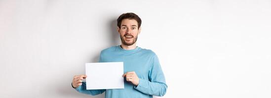 Romantic man looking with hopeful face at camera, showing sign card, blank piece of paper for banner or logo, standing over white background photo
