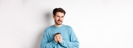 Handsome caucasian man warm-up hands with cup of coffee, smiling at camera and looking tenderly, standing on white background photo