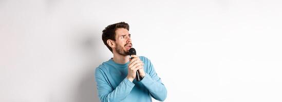 Worried and confused man reading lyrics on karaoke, looking left with unsure face, holding microphone and singing, white background photo