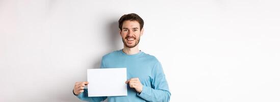 Young man with happy face showing blank piece of paper for your logo or sign, smiling at camera, standing over white background photo