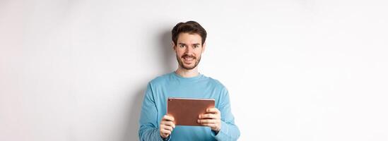 Handsome young man holding digital tablet and smiling, networking with device, standing over white background photo