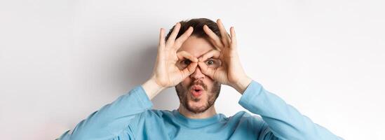 Impressed caucasian man looking through hand binoculars with amazed face, saying wow and checking out advertisement, white background photo