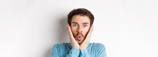 Close-up of impressed man checking out special offer, saying wow and touching face with amazed expression, stare at camera in awe, standing over white background photo