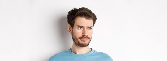 Close-up of disappointed sad man, grimacing and frowning, looking left upset, standing doubtful against white background photo