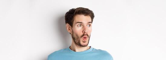 Close-up of impressed caucasian man saying wow, looking left amazed, checking out promotion deal, white background photo