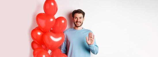 Handsome boyfriend saying hello, bring romantic red heart balloons on date, waving hand and smiling, standing over white background photo
