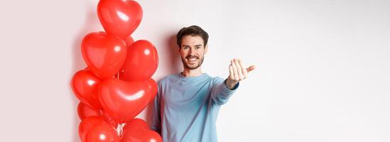 Smiling man beckon you to come closer, follow me gesture, taunting his lover move forward, have romantic surprise, standing near red balloon on valentines day, white background photo