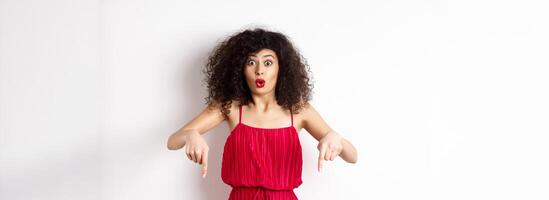 Impressed woman in red dress say wow, showing promo deal, pointing fingers down with amazed face, standing over white background photo