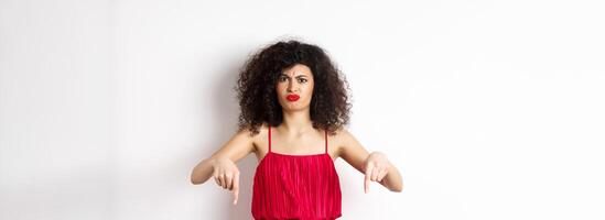 Disappointed caucasian woman in red dress complaining on spoiled date, frowning and grimacing, standing over white background photo