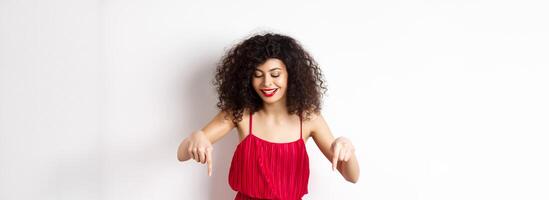 Cheerful elegant woman in red dress and makeup, looking and pointing down with pleased smile, showing advertisement, standing over white background photo