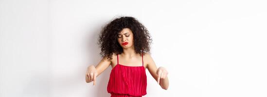 Sad and disappointed curly-haired woman in red dress, frowning and looking down jealous, pointing at promo, standing over white background photo