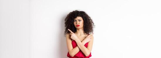 Doubtful and sad young woman in red dress, pointing fingers sideways and sulking, cant decide between products, need help with choice, standing over white background photo